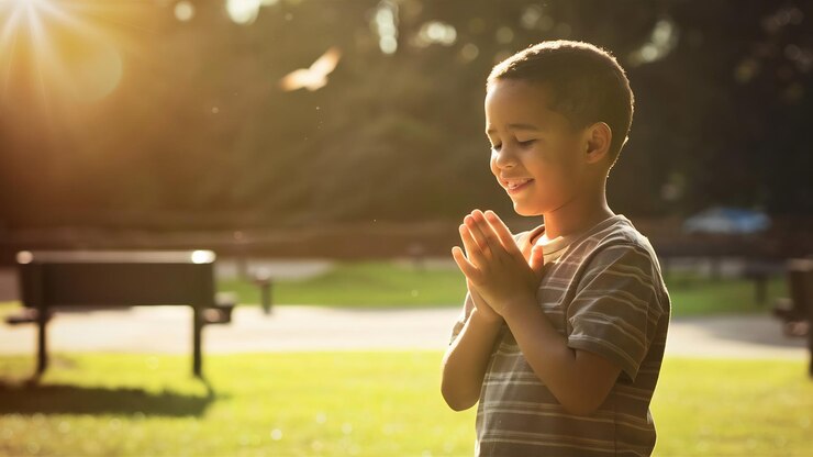 Foto menino de pé em um parque e orando sob a luz do sol com um fundo desfocado