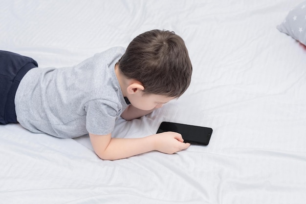 Menino deitado na cama com smartphone jogando ou estudando algo nele Conceito social e de tecnologia