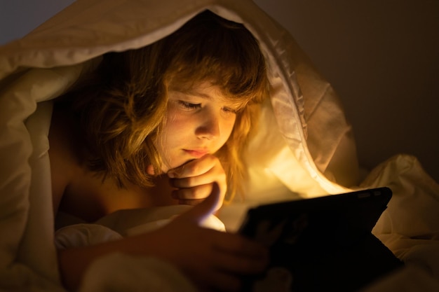 Menino deitado na cama e navegando na internet no tablet em um quarto escuro criança usando tablet PC à noite