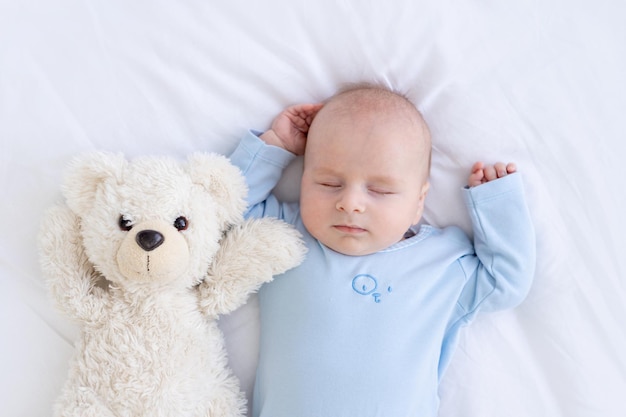 Menino dorme na cama deitado de costas com um urso de brinquedo macio de pijama azul, segurando as mãos para dormir saudável recém-nascido