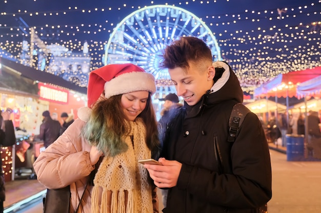 Menino e menina adolescentes no mercado de Natal, com chapéu de Papai Noel, com um smartphone