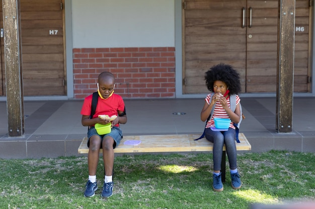 Menino e menina almoçando da caixa tiffin enquanto está sentado no banco do parque na escola