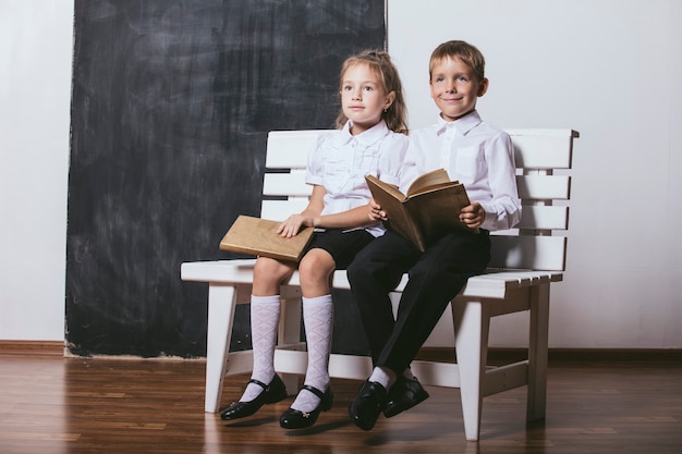 Menino e menina felizes da classe do ensino fundamental no banco lendo livros no fundo da lousa