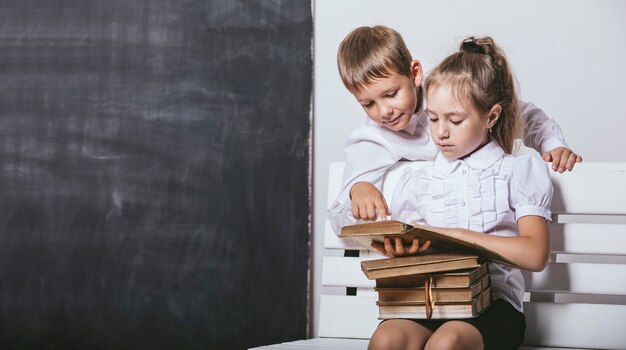 Menino e menina felizes da classe do ensino fundamental no banco lendo livros no fundo da lousa