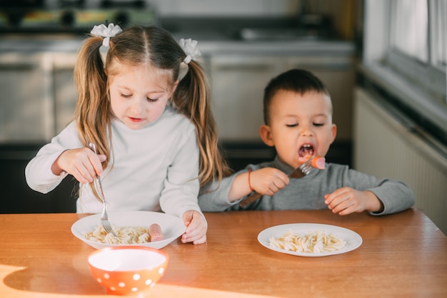 Menino e menina na cozinha comendo linguiça com macarrão é muito divertido e amigável