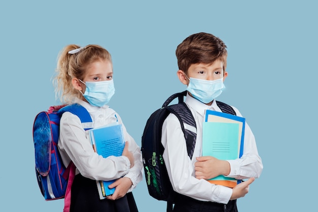 Menino e uma menina com máscara médica e mochila tem cadernos e livros na mão isolados em azul.