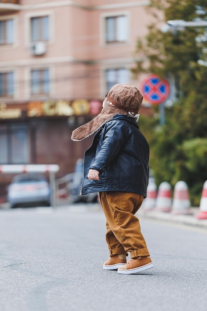 Menino elegante de 3 anos em uma jaqueta de couro e calça marrom anda na rua Criança moderna Moda infantil Criança feliz