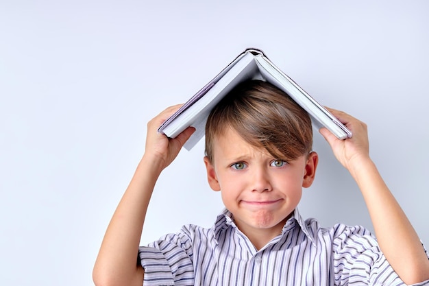 Menino em idade escolar com o livro na cabeça no espaço da cópia do fundo branco do estúdio.