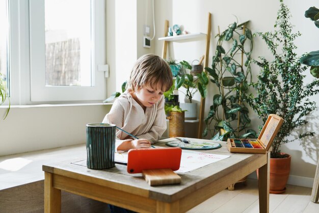 Menino em idade pré-escolar usando arte de tablet digital desenhando imagens multicoloridas usando tintas e pincel na mesa