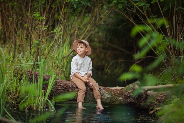 Menino em roupas rústicas e chapéu de vime senta-se com uma vara de pescar nas mãos e pega peixe num tronco do outro lado do rio. hobbies e lazer para homens.