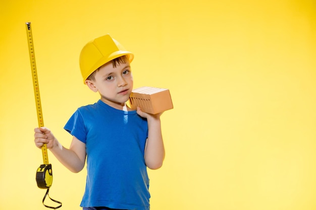 Menino em um capacete de construção segura um tijolo nas mãos e mede-o com uma fita métrica em estúdio em fundo amarelo