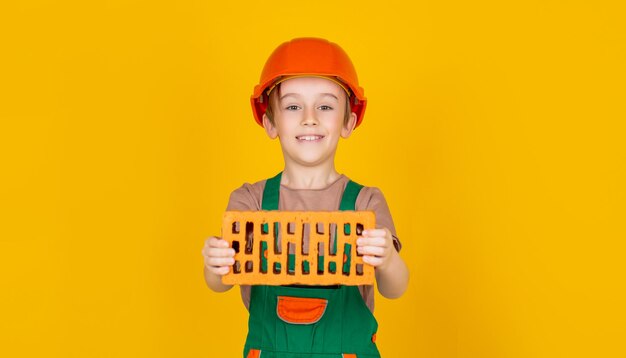 Menino em um capacete de construção tem um tijolo nas mãos em fundo amarelo Pequeno construtor de capacete Criança vestida como um construtor de operário Garotinho usando capacete Criança construindo capacete capacete