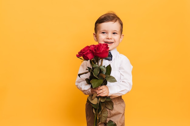 Menino em uma camisa branca, calça e gravata borboleta tem em suas mãos o buquê de rosas vermelhas. um presente para minha mãe