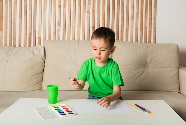 Menino em uma camiseta branca desenha com um pincel e pinta no papel em uma mesa na sala