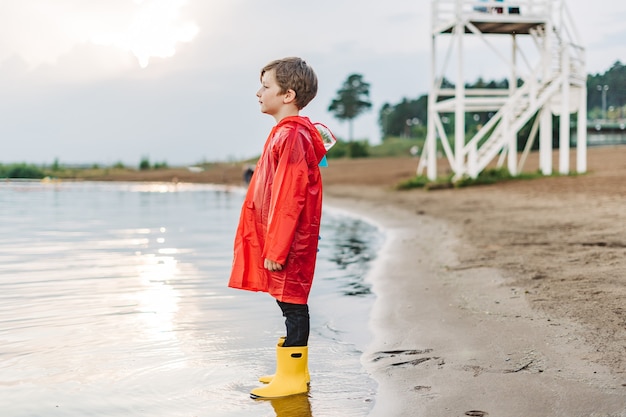 Menino em uma capa de chuva vermelha e botas de borracha amarelas brincando com água na praia.