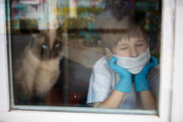 menino em uma máscara e luvas médicas, olhando pela janela com um gato