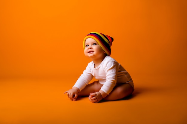 Foto menino em uma roupa branca e chapéu multicolorido, segurando um brinquedo sentado em uma parede amarela