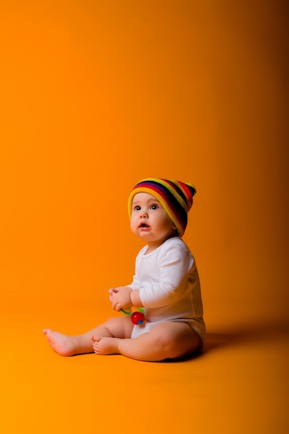 menino em uma roupa branca e chapéu multicolorido, segurando um brinquedo, sentado em uma parede laranja