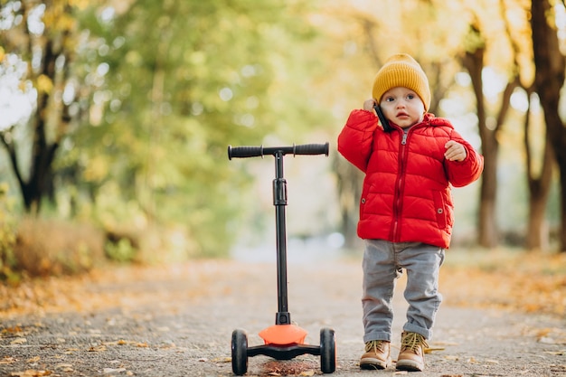 Menino em uma scooter segurando um telefone celular no parque outonal