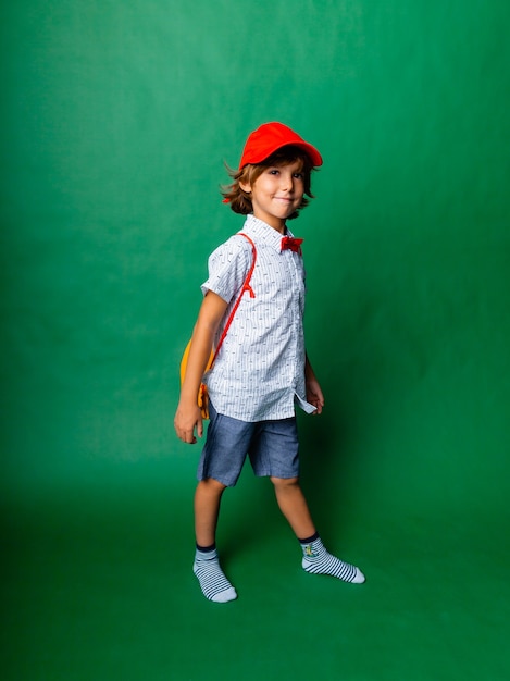 Menino emocional de 7 anos com um boné vermelho e uma mochila amarela. De pé sobre um fundo verde, se divertindo.