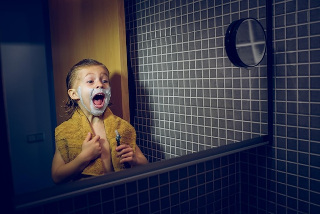 Menino encantado com espuma no rosto e lâmina de barbear na mão, olhando para o espelho com a boca aberta em pé no banheiro