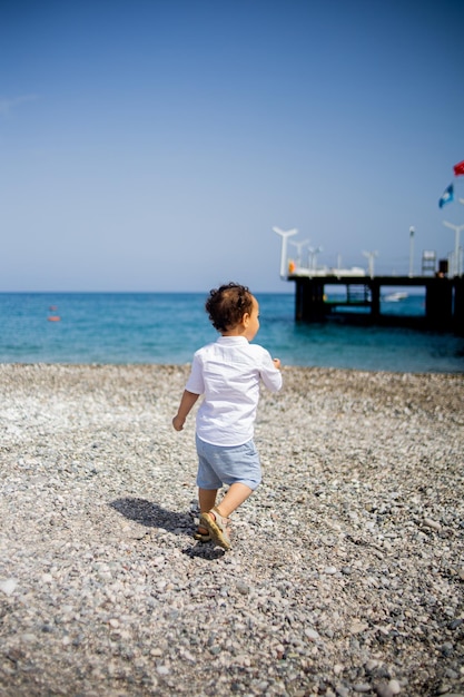 Menino encaracolado brinca na praia de calhau perto do mar azul