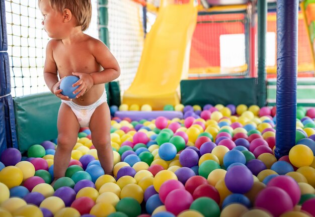 Menino engraçado alegre em uma fralda com uma bola listrada verde inflável nas mãos está jogando na sala de jogos no fim de semana desfrutando de dias quentes de verão brilhantes