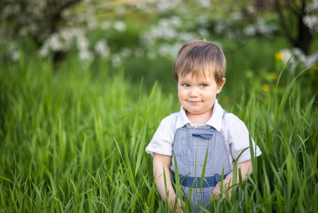 Menino engraçado com olhos azuis brilhantes de macacão comendo grama verde fresca em um grande jardim florido