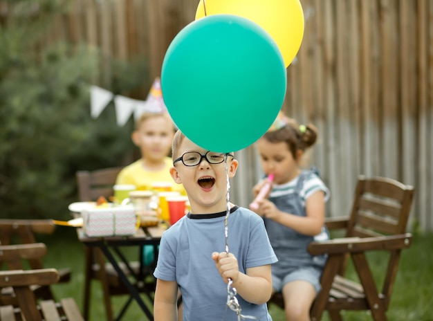 Menino engraçado de quatro anos comemorando seu aniversário com a família ou amigos em uma festa de aniversário no quintal garoto usando chapéu de festa e segurando balão na mão