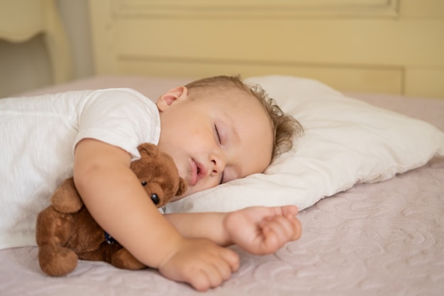 Menino engraçado dormindo na cama em casa. criança abraçando o ursinho de pelúcia.