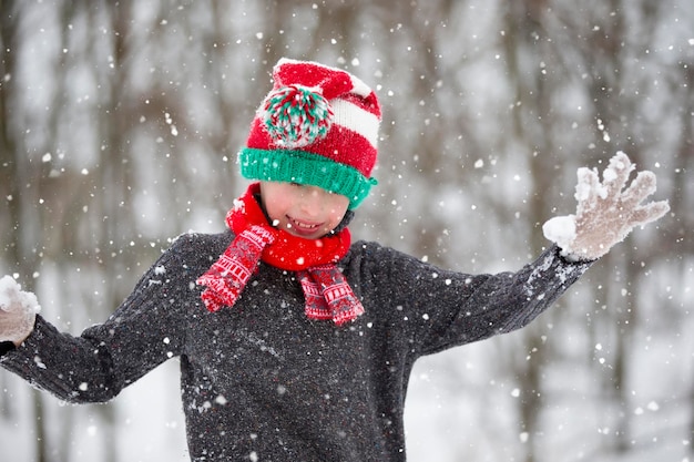 Menino engraçado em roupas de inverno caminha durante uma queda de neve Atividades ao ar livre de inverno para crianças