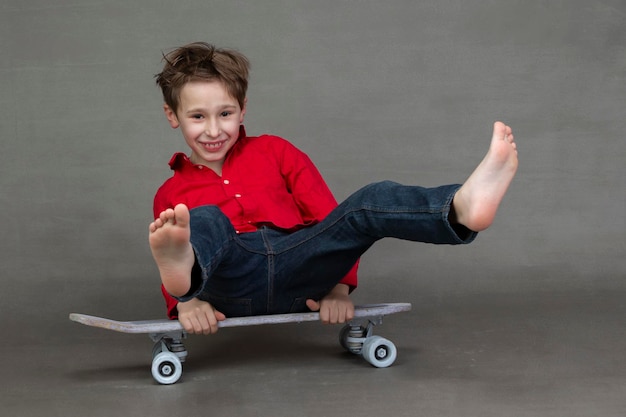 Menino engraçado sentado em um skate vintage com os pés descalços em um fundo cinza.