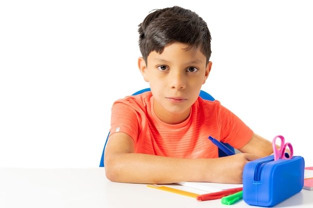 Menino escrevendo em seu caderno na mesa branca com vários lápis de cor e o estojo