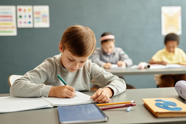 Menino escrevendo no caderno na secretária na sala de aula na escola