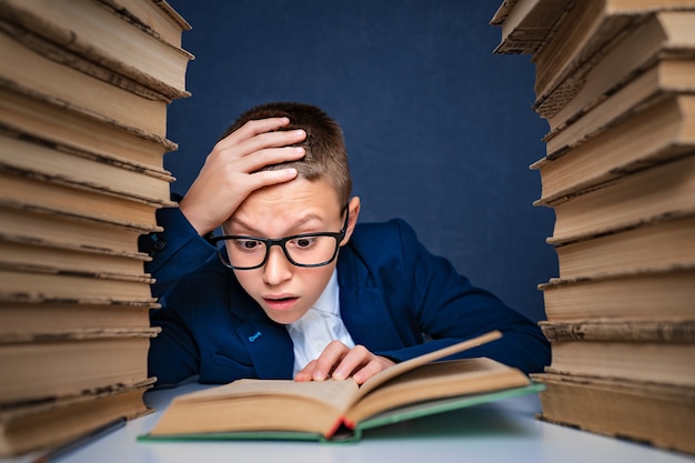 Menino esperto em copos sentado entre duas pilhas de livros e ler o livro pensativamente.