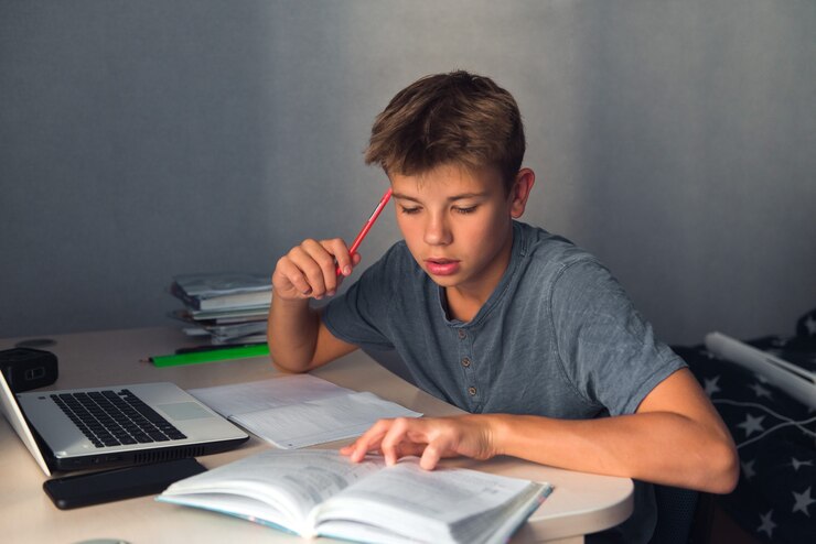 Menino estudante adolescente fazendo lição de casa com o caderno aberto do laptop e o computador no local de trabalho em casa
