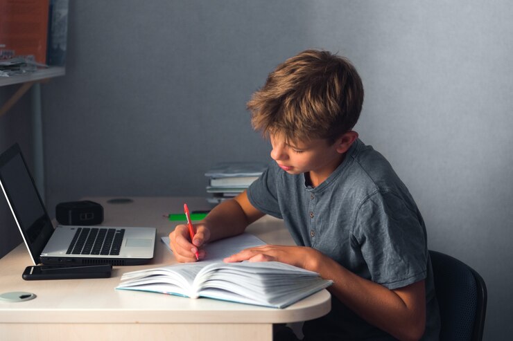Menino estudante adolescente fazendo lição de casa com o caderno aberto do laptop e o computador no local de trabalho em casa
