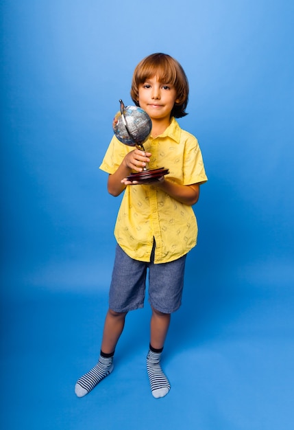 Menino estudante estudante de 7 anos com um globo de lembrança nas mãos sorrindo sobre um fundo azul