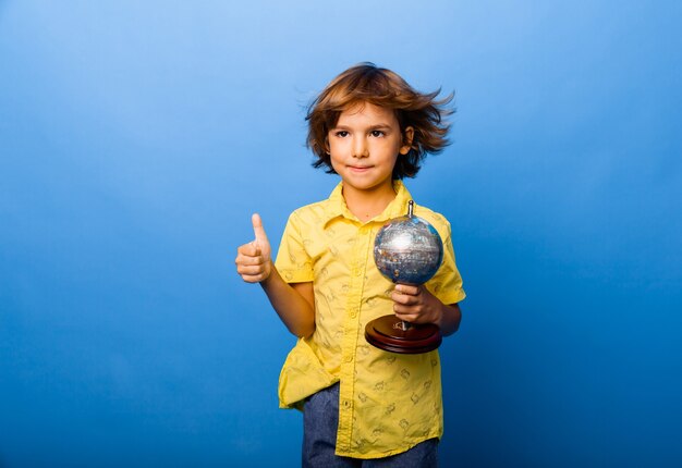 Menino estudante estudante de 7 anos com um globo de lembrança nas mãos sorrindo sobre um fundo azul