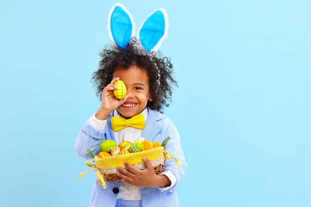 Foto menino étnico alegre com cabelo afro em fantasia de páscoa e orelhas de coelho sorrindo enquanto cobria o olho com ovo colorido contra fundo azul