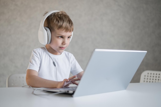 Menino europeu bonito ouve música em fones de ouvido e digita em um laptop A criança faz lição de casa no computador Um jovem estudante recebe educação a distância
