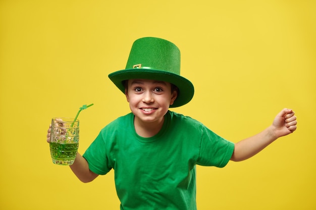 Menino feliz com chapéu de duende com as mãos para cima segura um copo com bebida verde e expressa a felicidade em comemoração ao dia de São Patrício