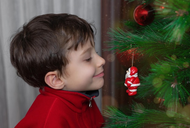 Menino feliz de close-up em uma jaqueta vermelha, olhando com curiosidade para um brinquedo de árvore de Natal.