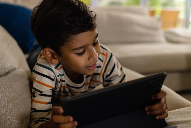 Menino feliz de duas raças deitado no sofá usando um tablet na sala de estar. Infância, tempo livre, tecnologia, comunicação, entretenimento, estilo de vida e vida doméstica inalterados.