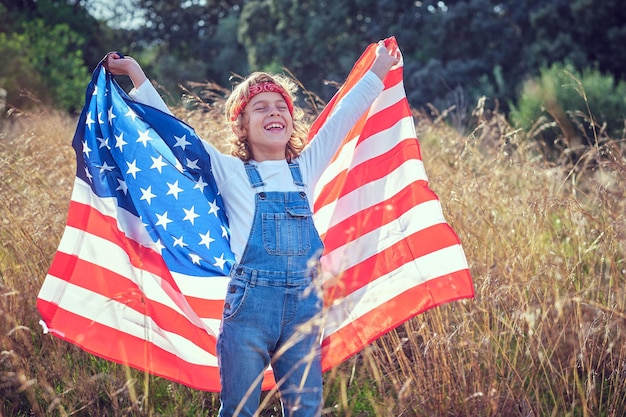 menino feliz em denim geral e com bandeira americana na mão sorrindo alegremente enquanto estava de pé em um campo gramado