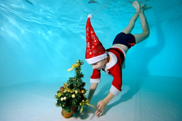 Menino feliz em um boné Papai Noel mergulha no fundo da piscina para pegar uma árvore de Natal