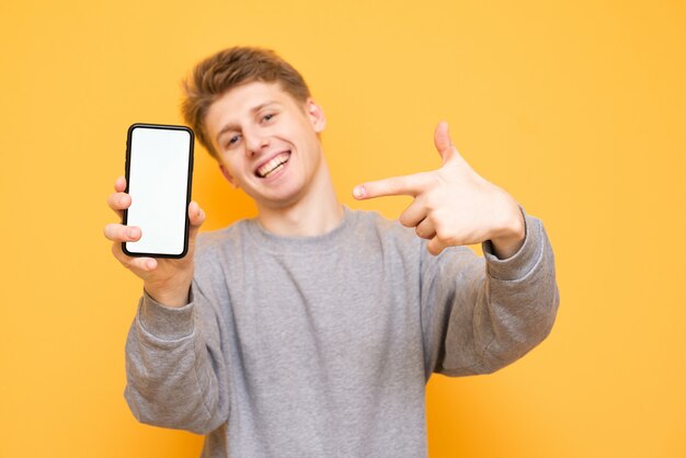 Menino feliz está de pé sobre um fundo amarelo, olha para a câmera e mostra os dedos no smartphone segura na mão