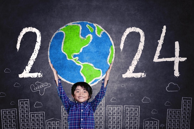Menino feliz levantando um globo desenhado com números de 2024 no quadro-negro em pé na frente do quadro-negro