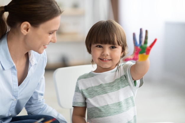 Menino feliz mostrando a mão colorida pintada e sorrindo para a câmera sentada na escola com tutor