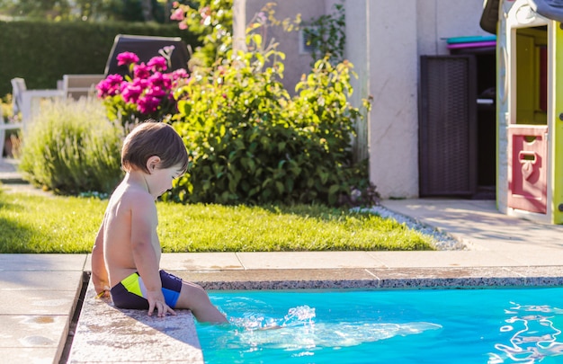 Foto menino feliz pequeno que senta-se no lado da piscina no jardim que joga por seus pés na água que tem o divertimento.